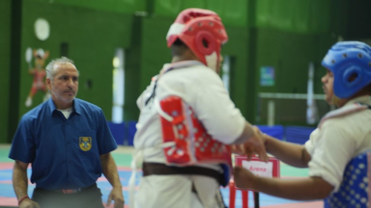 A referee guides a young fighter in a red helmet at a KKFI tournament